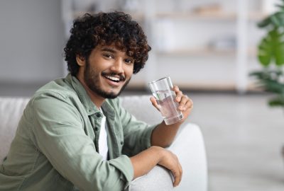 Handsome dark-skinned young man drinking water and smiling at camera, cheerful indian guy sitting on couch in living room, holding glass of fresh water, chilling alone at home, copy space